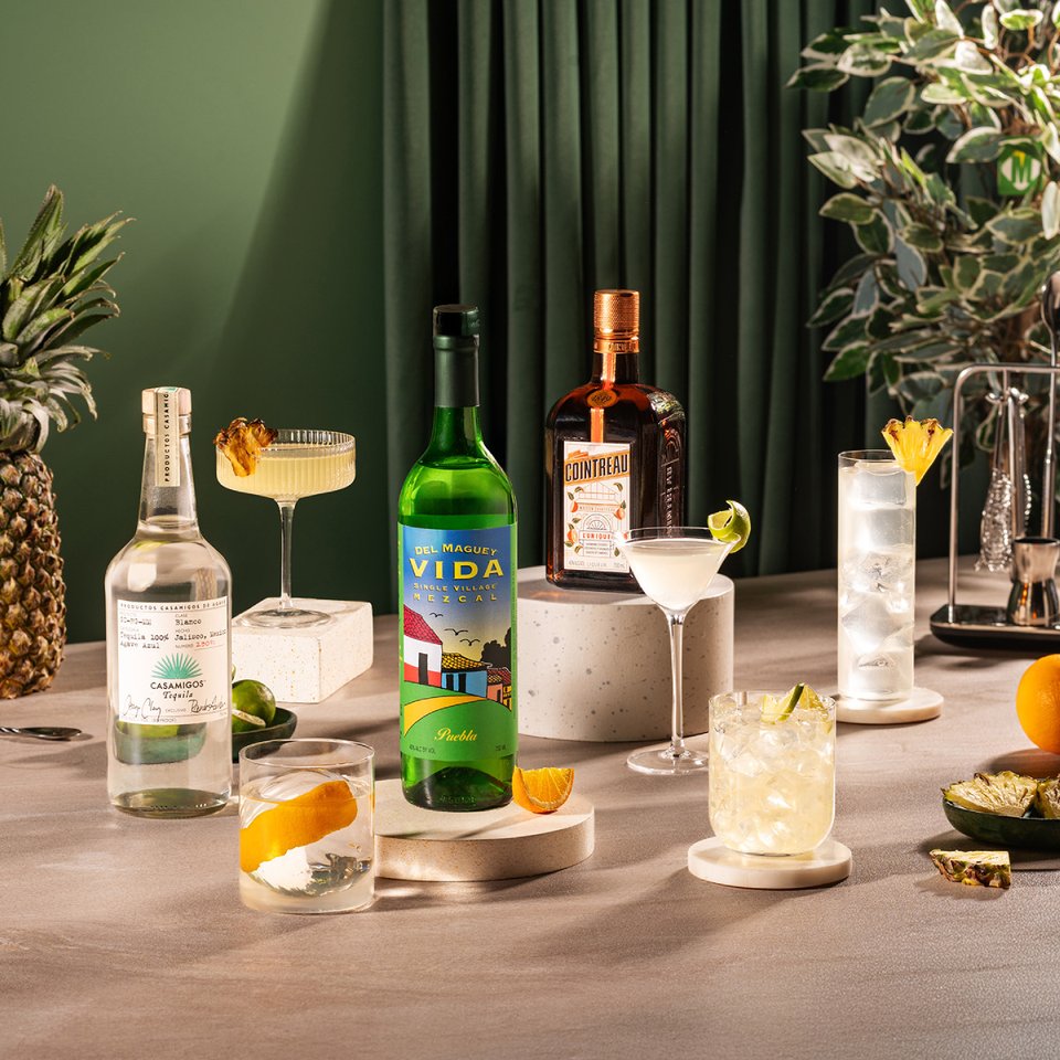 The Agave Lover's official cover: agave cocktails, pineapple and citrus beside tequila and mezcal bottles against green curtains.