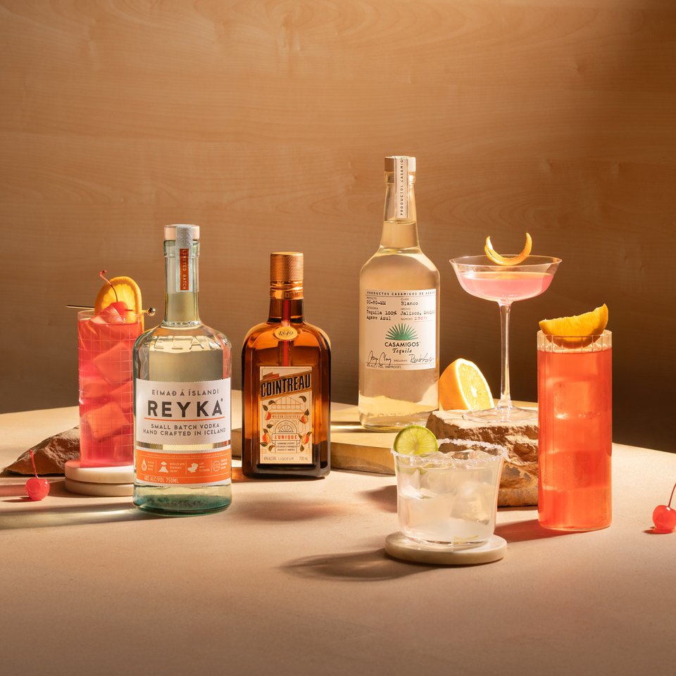 The Signature Mixlist: four citrus and cranberry cocktails with Reyka, Cointreau and Casamigos bottles on a warm wood backdrop.